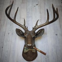 Faux taxidermy deer head painted rubbed bronze with rubbed bronze antlers and gold accents. A cigar is in the deers mouth and a bow tie accents the deers neck.
