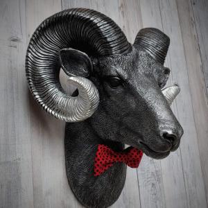 Faux taxidermy ram head wall mount, painted black with metallic silver tipped horns with graduating silver going to the head. A red and black polka dot bow tie accents the neck of the ram.