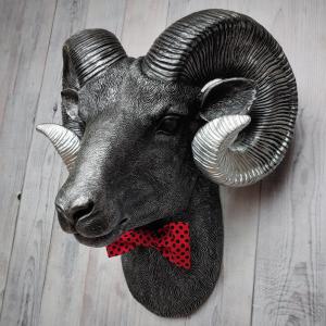 Faux taxidermy ram head wall mount, painted black with metallic silver tipped horns with graduating silver going to the head. A red and black polka dot bow tie accents the neck of the ram.