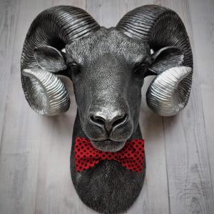 Faux taxidermy ram head wall mount, painted black with metallic silver tipped horns with graduating silver going to the head. A red and black polka dot bow tie accents the neck of the ram.