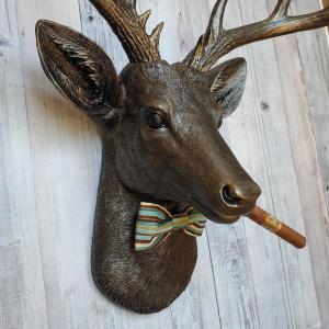 Faux taxidermy deer head painted rubbed bronze with rubbed bronze antlers and gold accents. A cigar is in the deers mouth and a bow tie accents the deers neck.