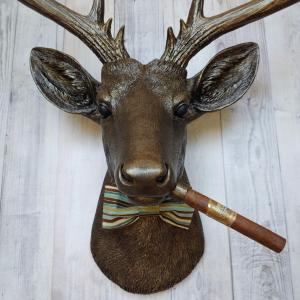 Faux taxidermy deer head painted rubbed bronze with rubbed bronze antlers and gold accents. A cigar is in the deers mouth and a bow tie accents the deers neck.