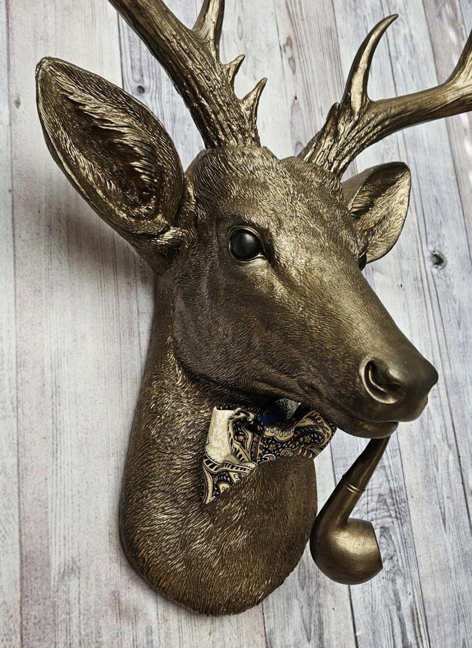 Faux taxidermy deer head with smoking pipe and bow tie, painted antique gold with dark bronze accents.