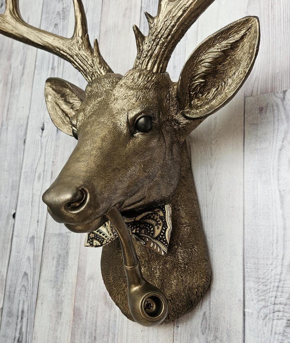 Faux taxidermy deer head with smoking pipe and bow tie, painted antique gold with dark bronze accents.