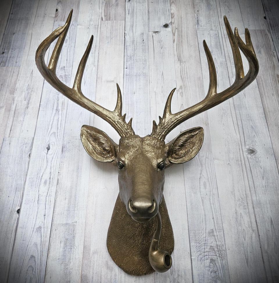 Faux taxidermy deer head wall mount with smoking pipe, painted antique gold with dark bronze accenting.