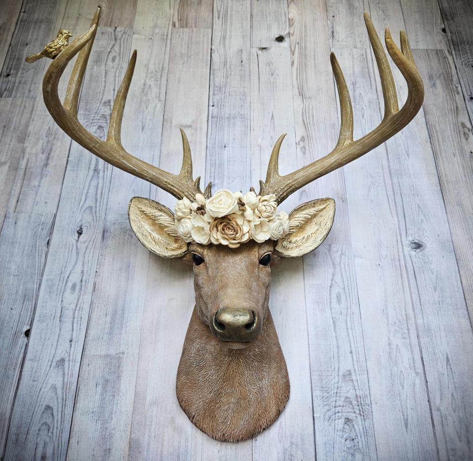Faux taxidermy deer head, painted in natural colors with a brass bird on one antler and a wooden flower crown atop it's head.