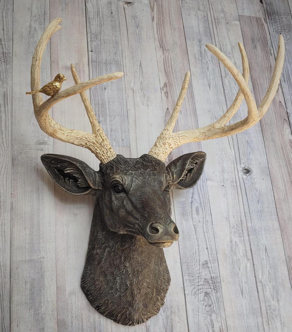 Faux taxidermy deer head painted chestnut brown with cream and gold accenting. Cream antlers with gold antlers, a brass bird rests atop one of the antlers.