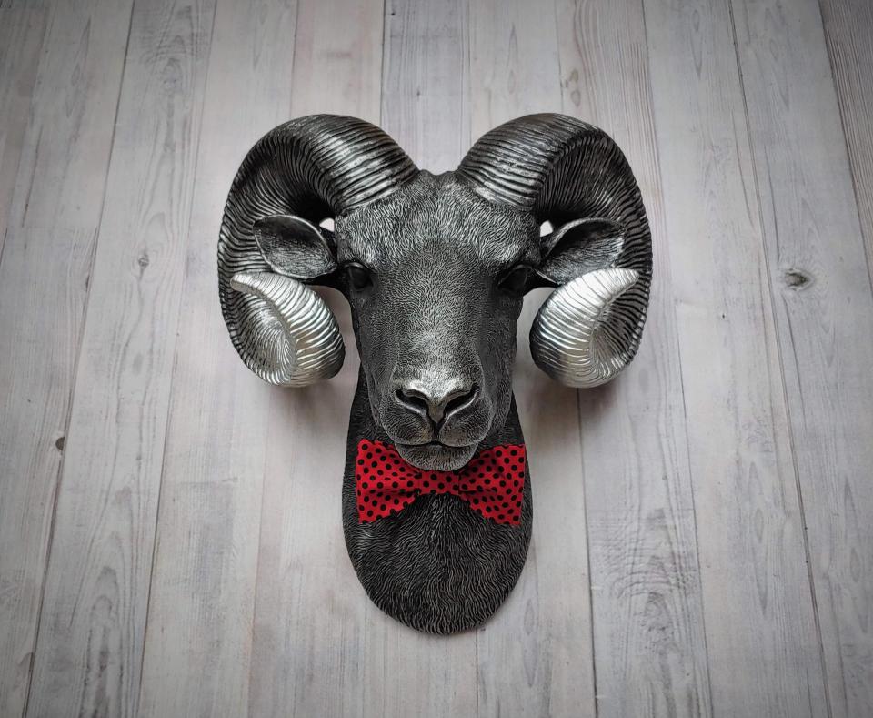 Faux taxidermy ram head wall mount, painted black with metallic silver tipped horns with graduating silver going to the head. A red and black polka dot bow tie accents the neck of the ram.