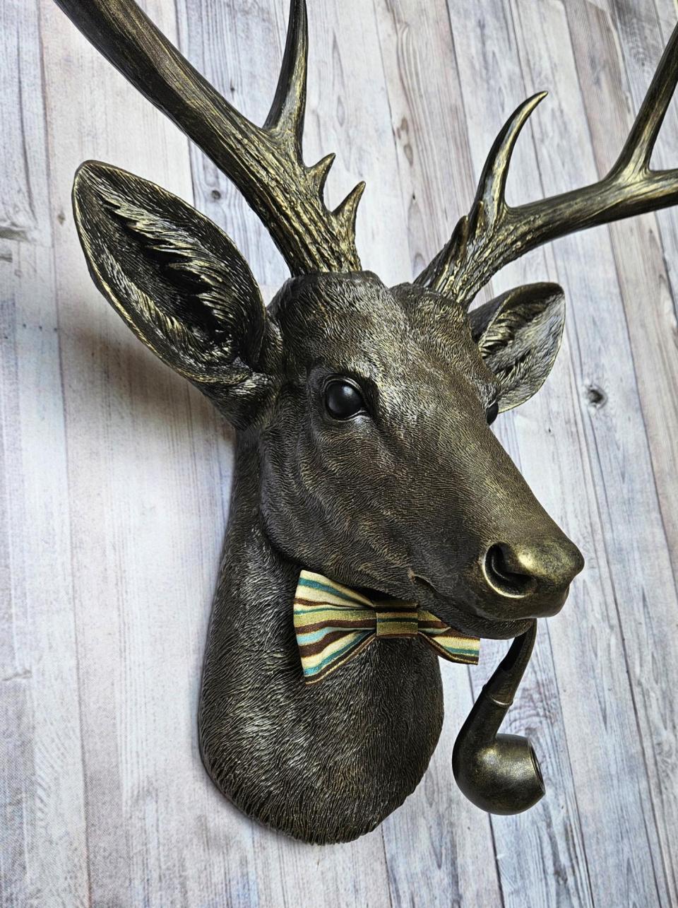 faux taxidermy deer head, rubbed bronze with gold accents. A smoking pipe hangs from the deers mouth and its wearing a striped bow tie