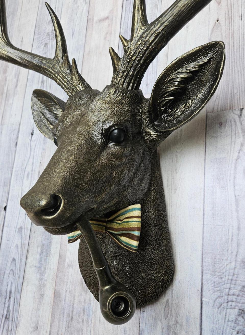 faux taxidermy deer head, rubbed bronze with gold accents. A smoking pipe hangs from the deers mouth and its wearing a striped bow tie