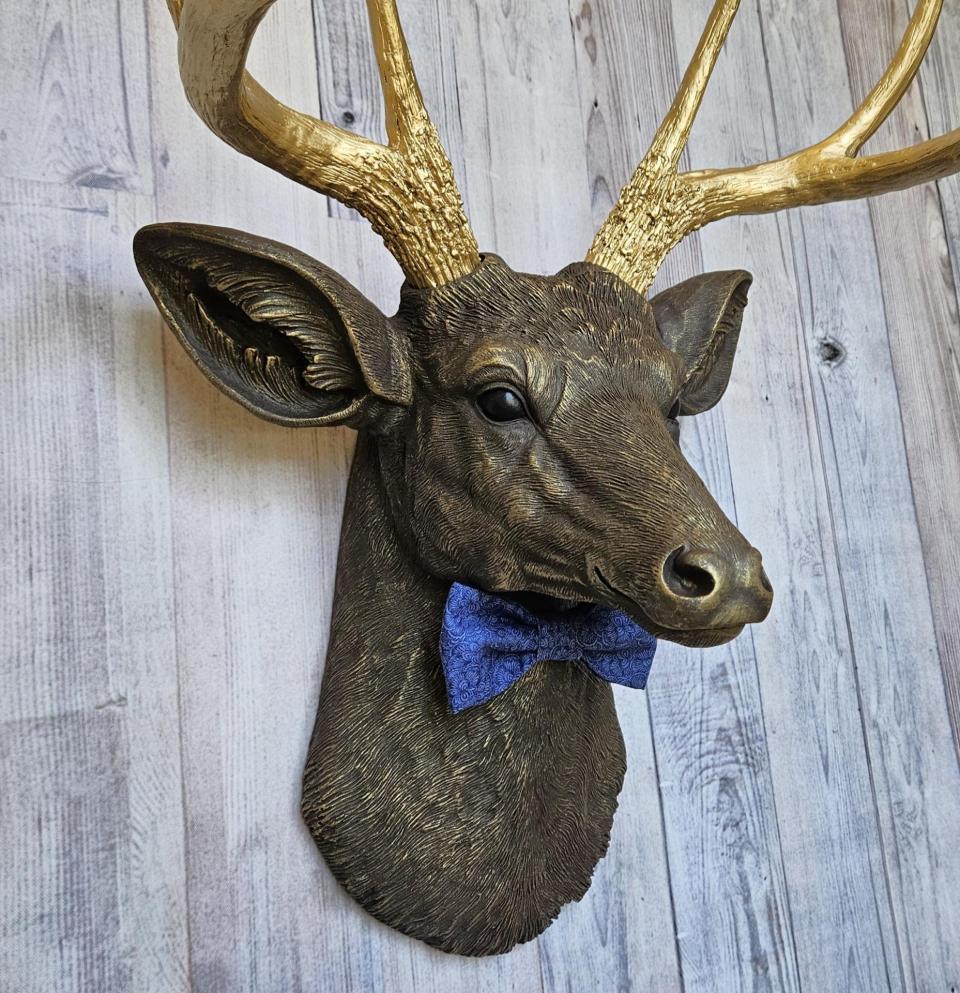 Faux taxidermy deer head painted chestnut brown with gold antlers. A blue bow tie accents the deers neck.