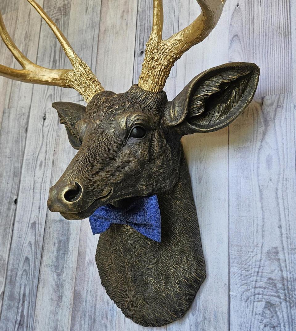 Faux taxidermy deer head painted chestnut brown with gold antlers. A blue bow tie accents the deers neck.