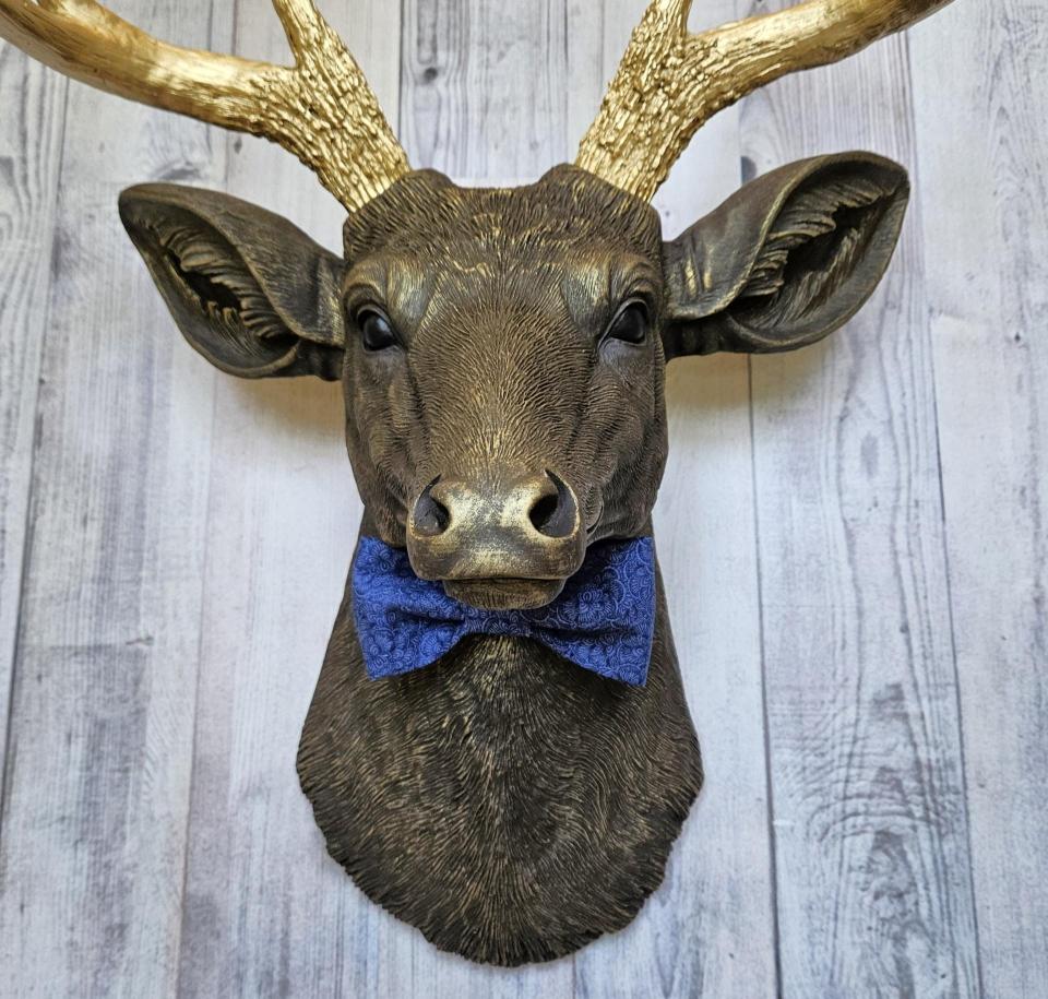 Faux taxidermy deer head painted chestnut brown with gold antlers. A blue bow tie accents the deers neck.