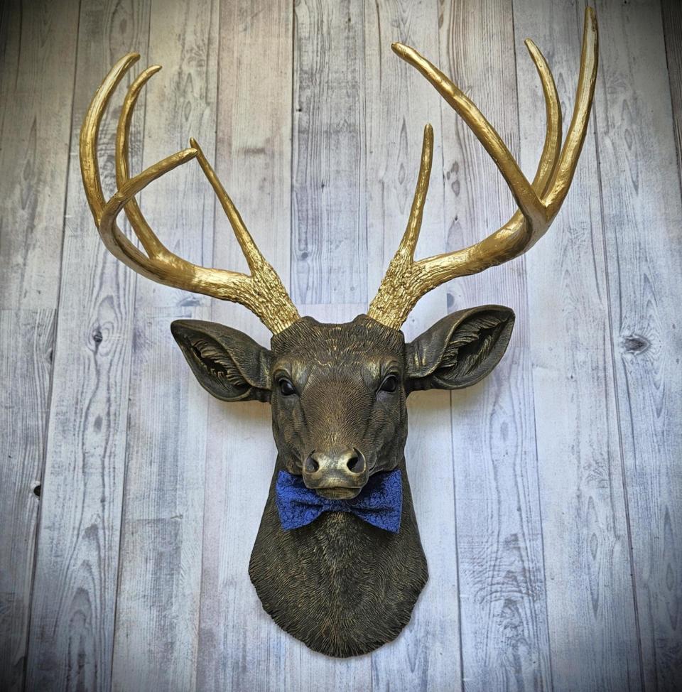 Faux taxidermy deer head painted chestnut brown with gold antlers. A blue bow tie accents the deers neck.