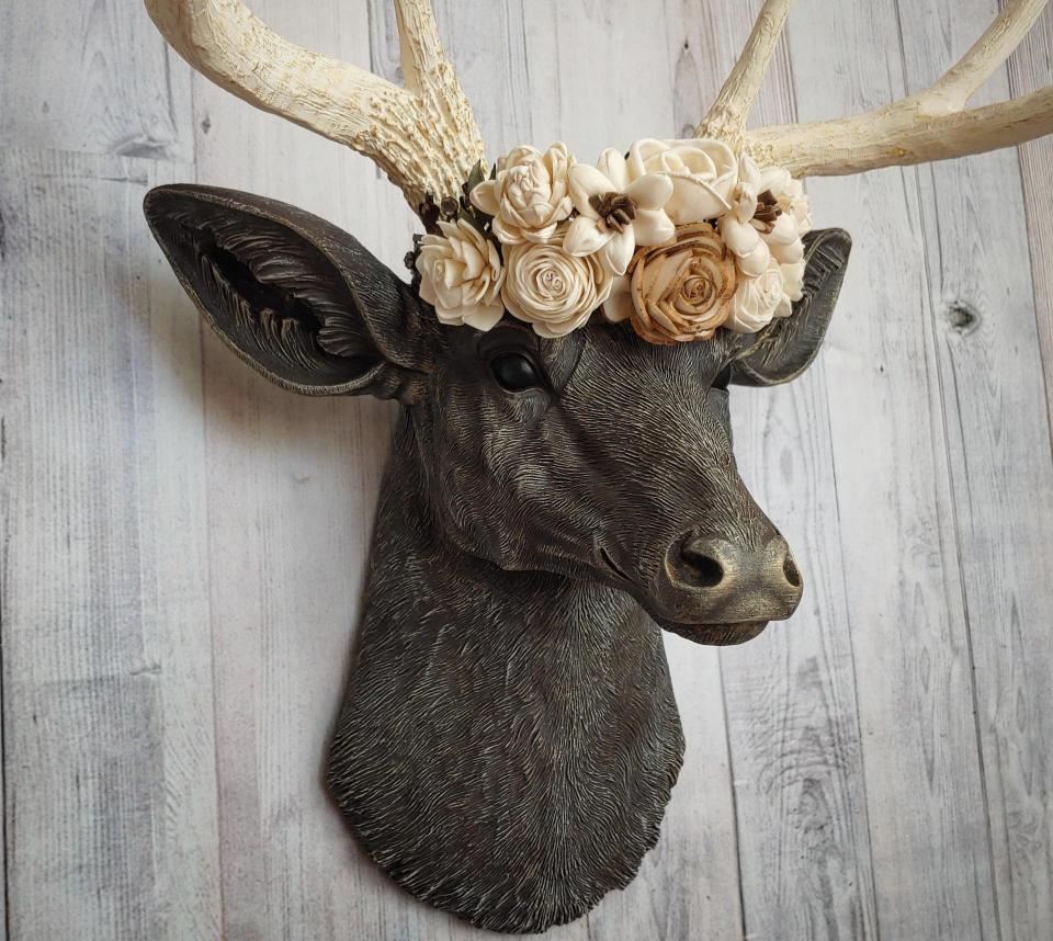 Faux taxidermy deer head, painted chestnut brown with antique white and gold accenting. Antlers are antique white with gold and chestnut brown accenting. A cream floral crown rests atop the deers head.