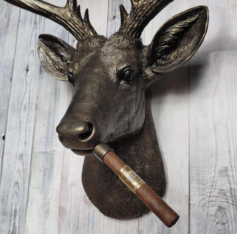 Faux taxidermy deer head painted rubbed bronze with rubbed bronze antlers and gold accents. A cigar is in the deers mouth.