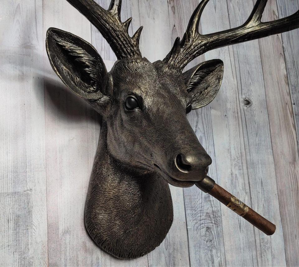 Faux taxidermy deer head painted rubbed bronze with rubbed bronze antlers and gold accents. A cigar is in the deers mouth.