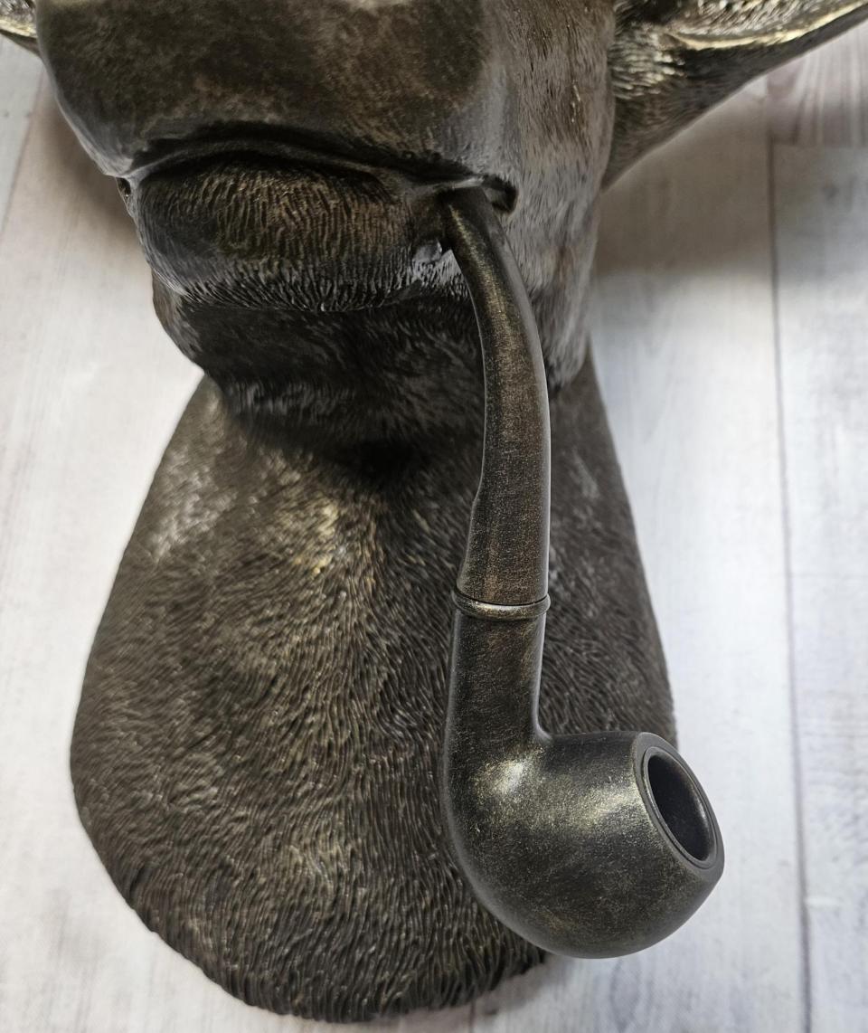 Rubbed bronze faux taxidermy deer head with gold accents. A smoking pipe extends from the deers mouth