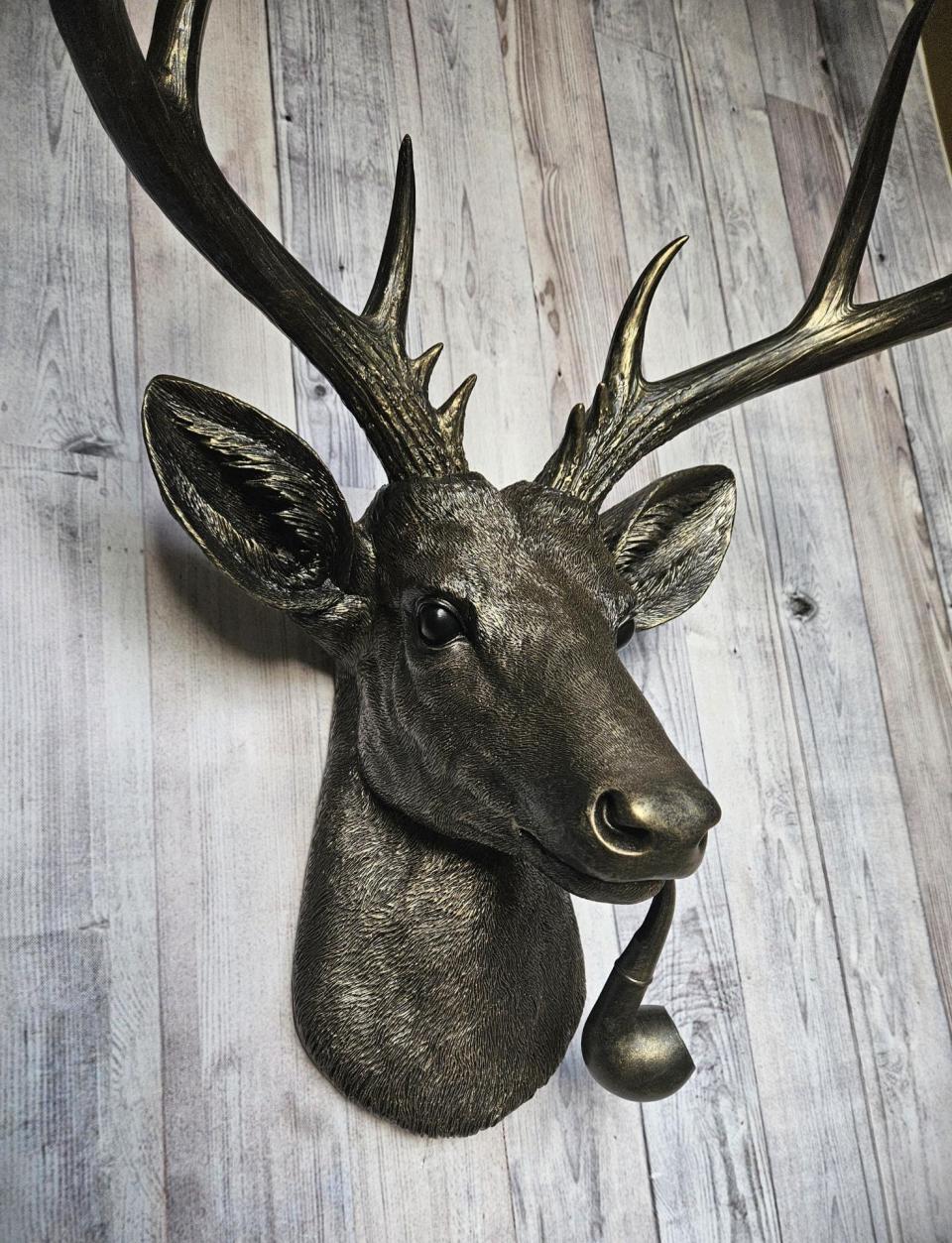 Rubbed bronze faux taxidermy deer head with gold accents. A smoking pipe extends from the deers mouth