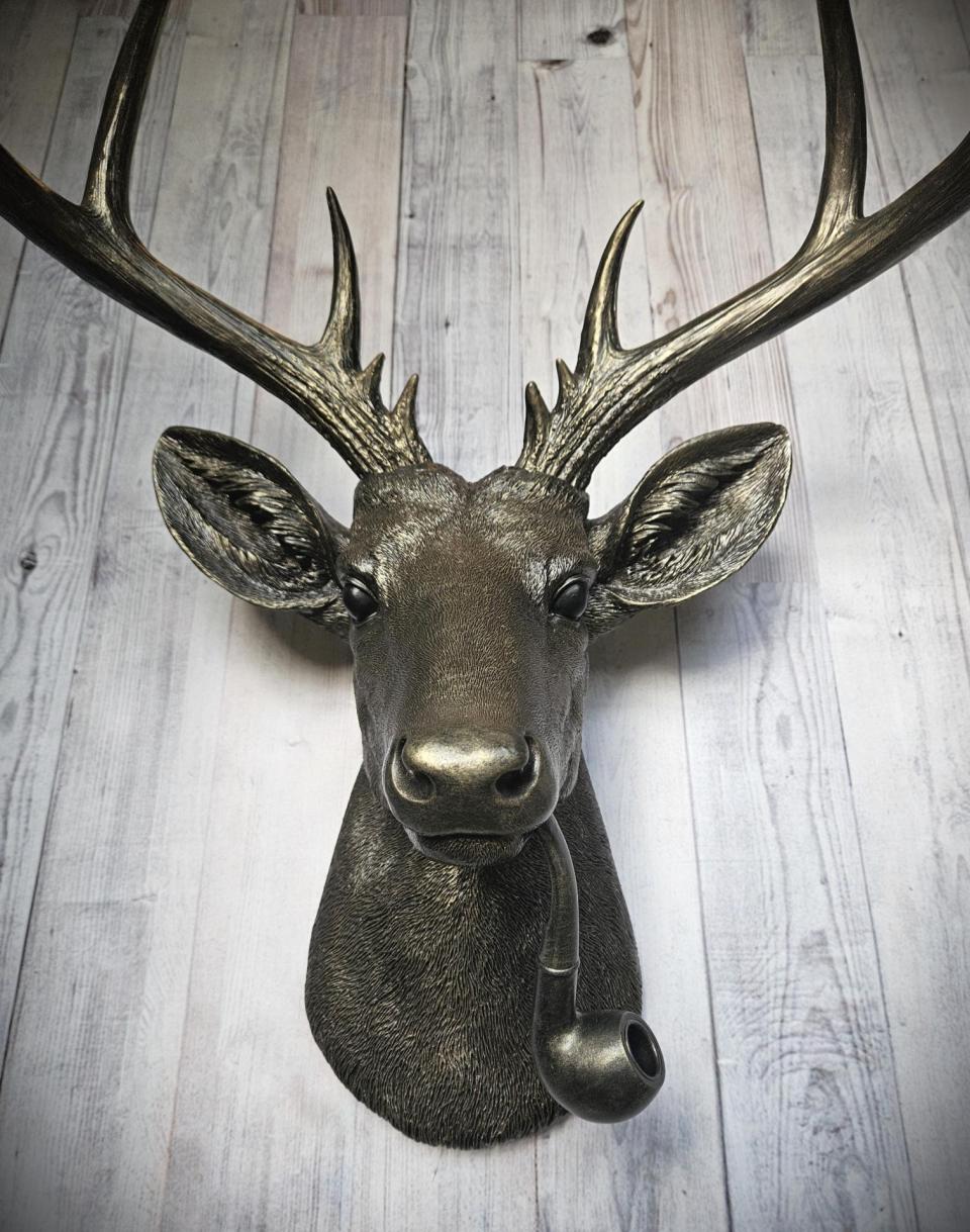 Rubbed bronze faux taxidermy deer head with gold accents. A smoking pipe extends from the deers mouth