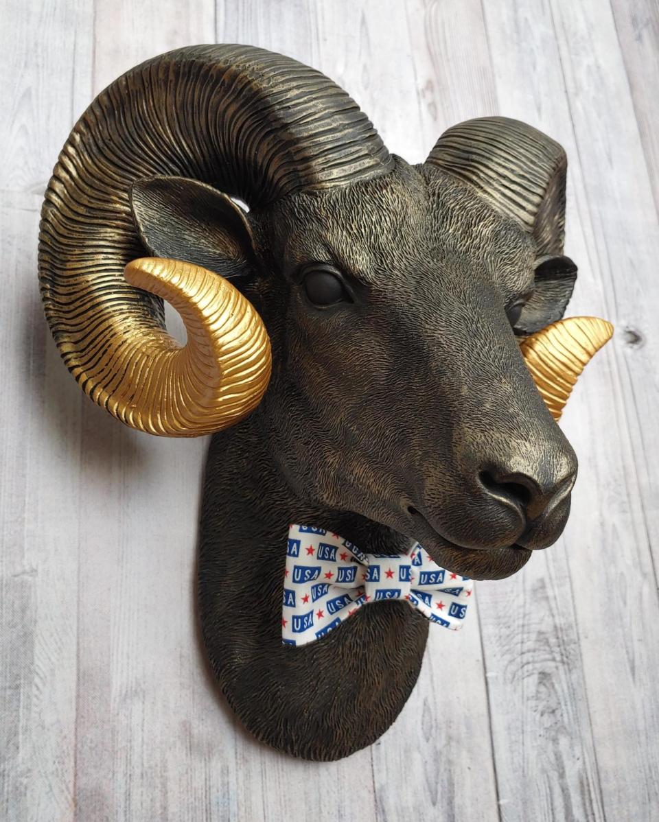 Faux taxidermy ram head wall mount, painted black with metallic gold tipped horns with graduating gold going to the head. A USA bow tie accents the neck of the ram.