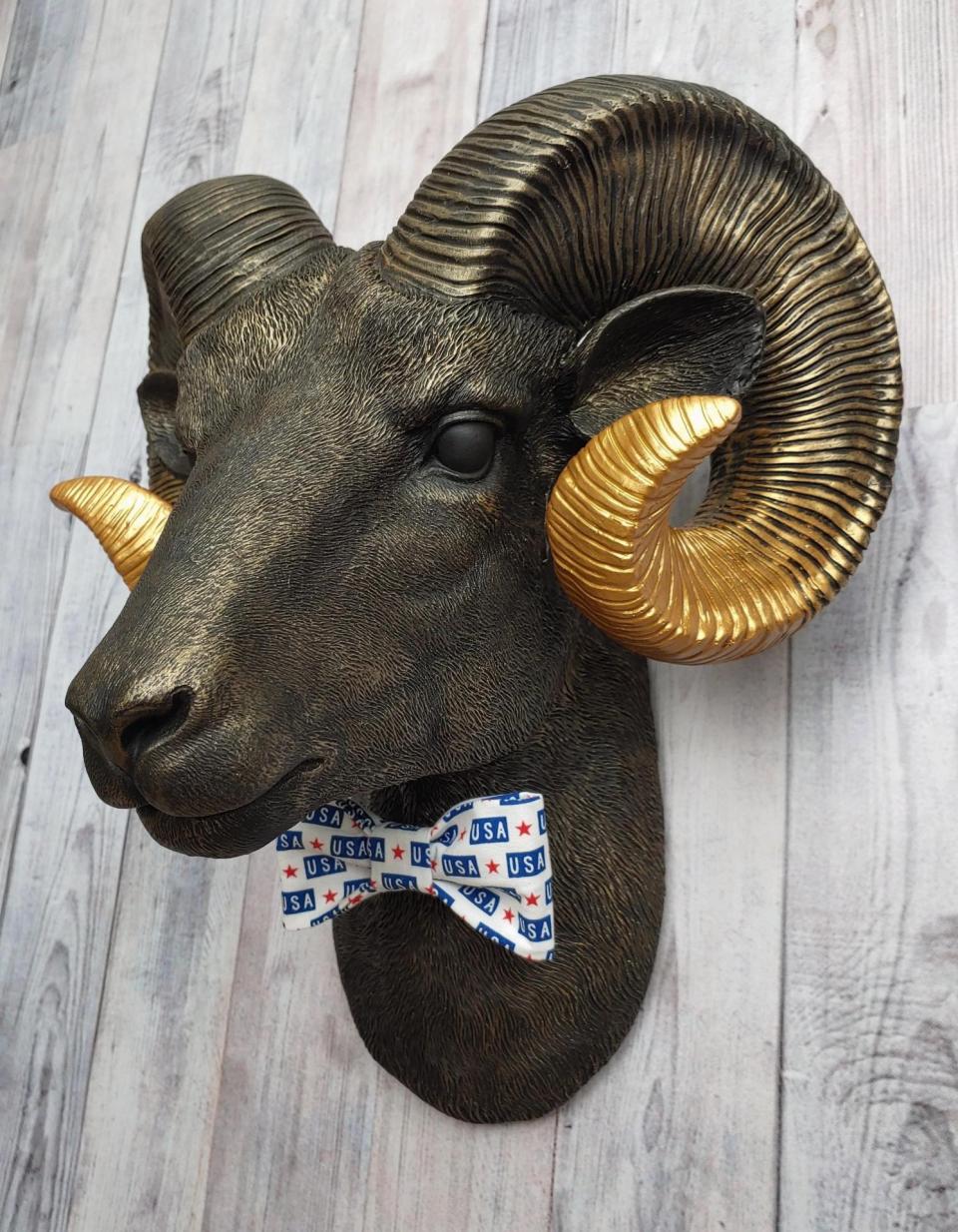 Faux taxidermy ram head wall mount, painted black with metallic gold tipped horns with graduating gold going to the head. A USA bow tie accents the neck of the ram.