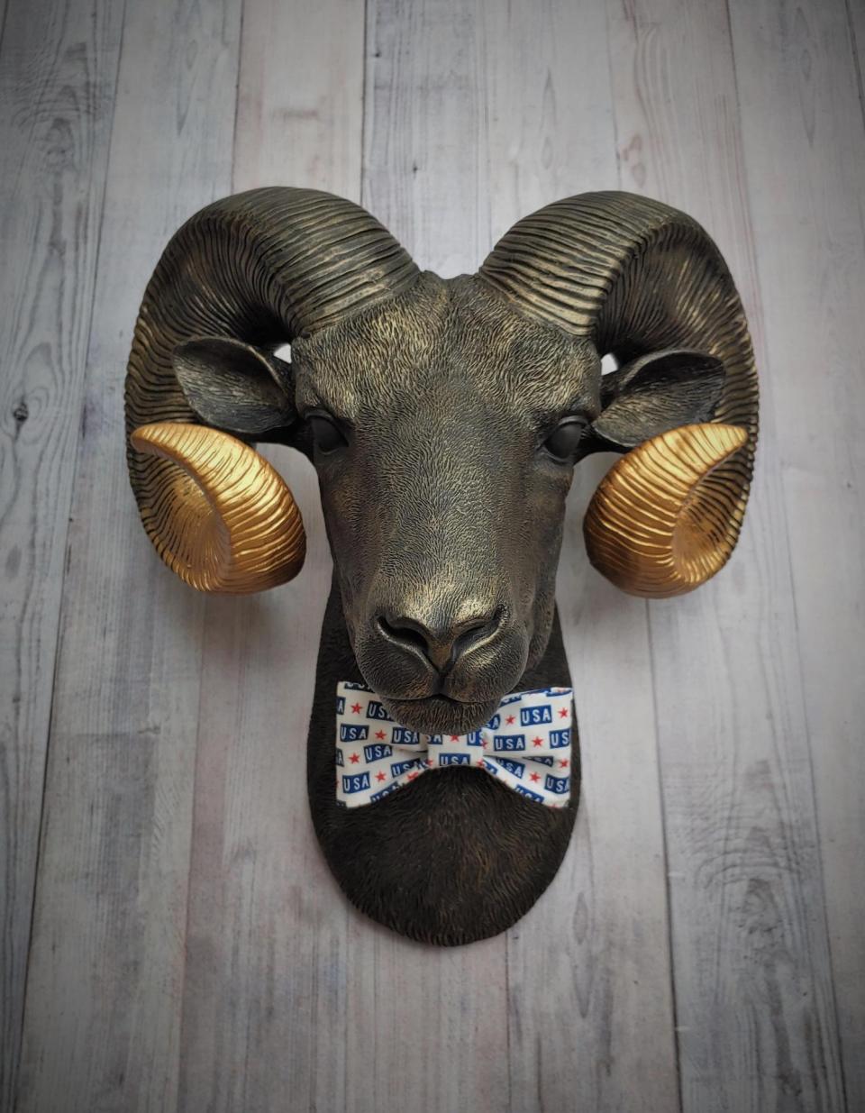 Faux taxidermy ram head wall mount, painted black with metallic gold tipped horns with graduating gold going to the head. A USA bow tie accents the neck of the ram.