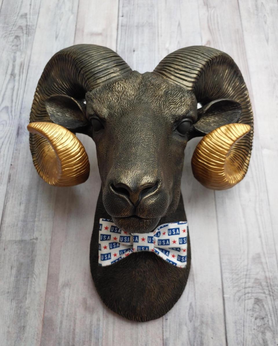 Faux taxidermy ram head wall mount, painted black with metallic gold tipped horns with graduating gold going to the head. A USA bow tie accents the neck of the ram.