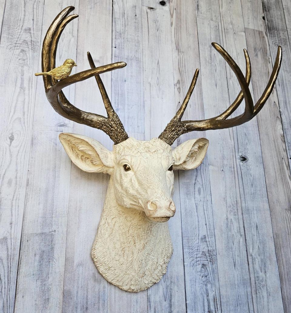 Faux taxidermy deer head, painted antique white with antique gold accenting. The antlers are antique gold a small brass bird is perched on the left facing antler.