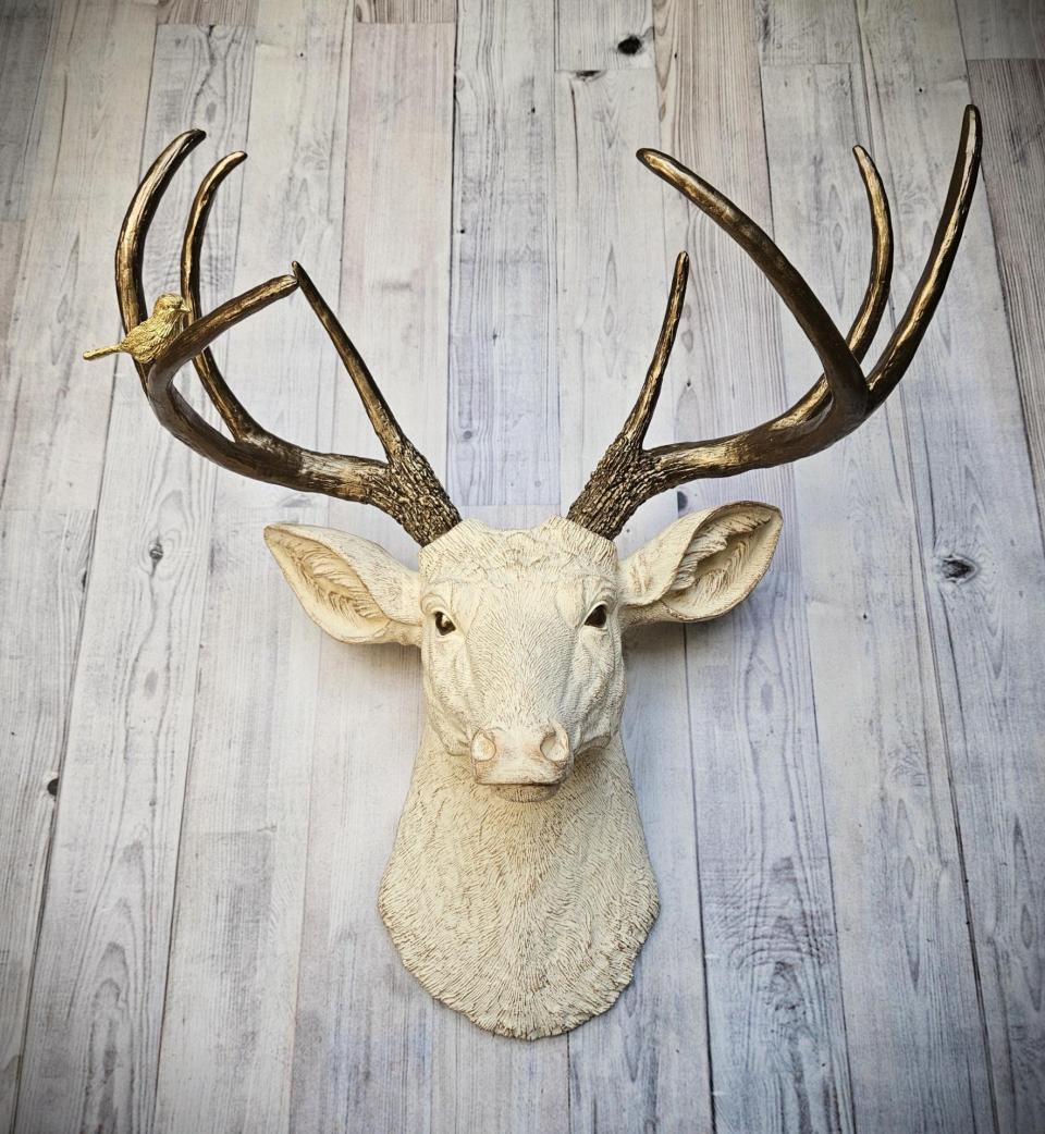 Faux taxidermy deer head, painted antique white with antique gold accenting. The antlers are antique gold a small brass bird is perched on the left facing antler.