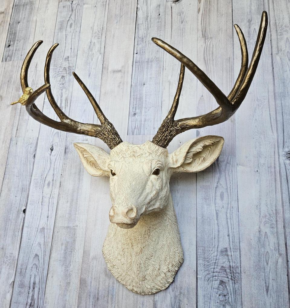 Faux taxidermy deer head, painted antique white with antique gold accenting. The antlers are antique gold a small brass bird is perched on the left facing antler.