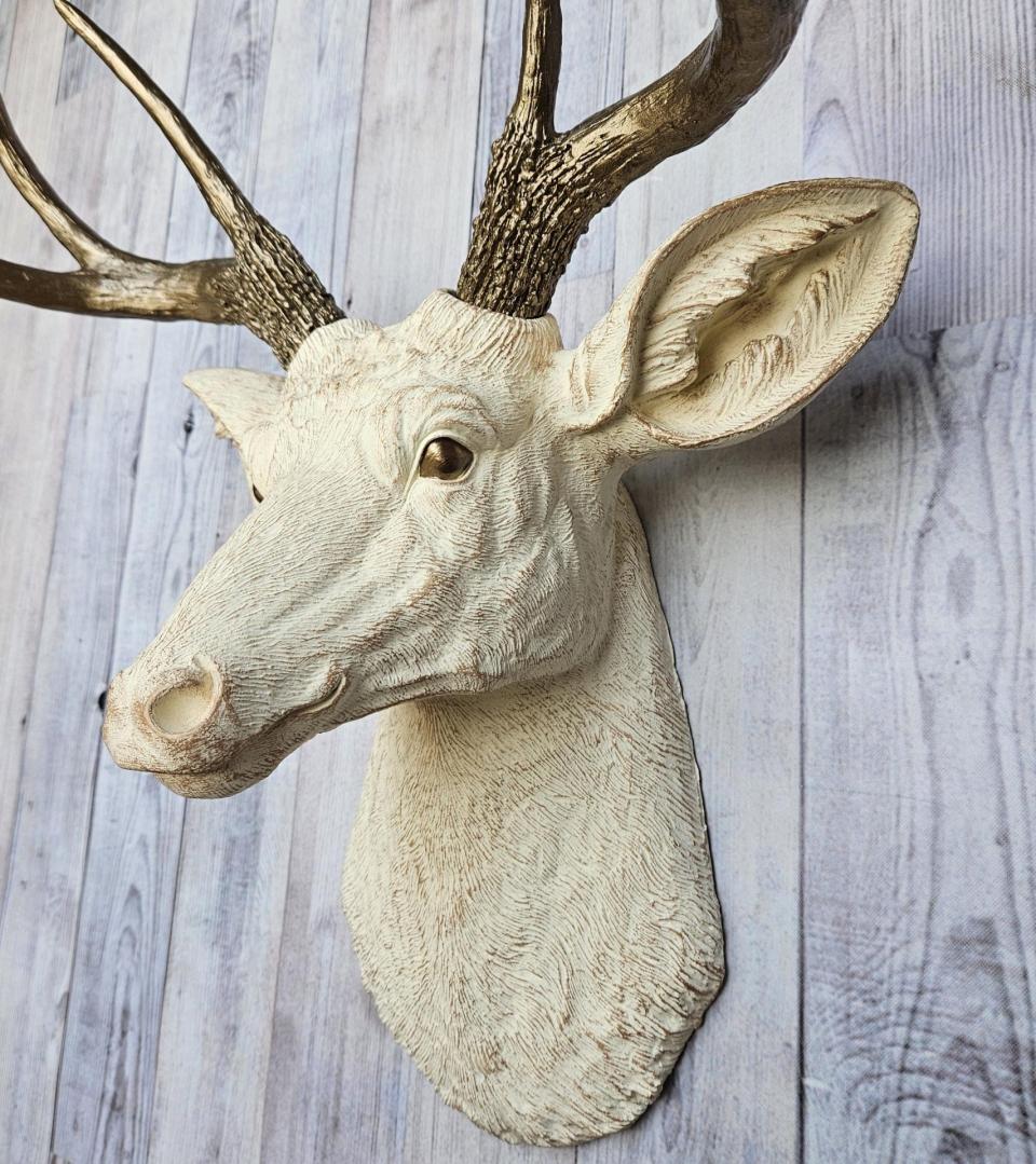 Faux taxidermy deer head, painted antique white with antique gold accenting. The antlers are antique gold a small brass bird is perched on the left facing antler.