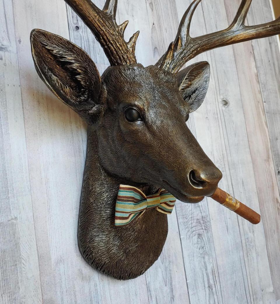 Faux taxidermy deer head painted rubbed bronze with rubbed bronze antlers and gold accents. A cigar is in the deers mouth and a bow tie accents the deers neck.