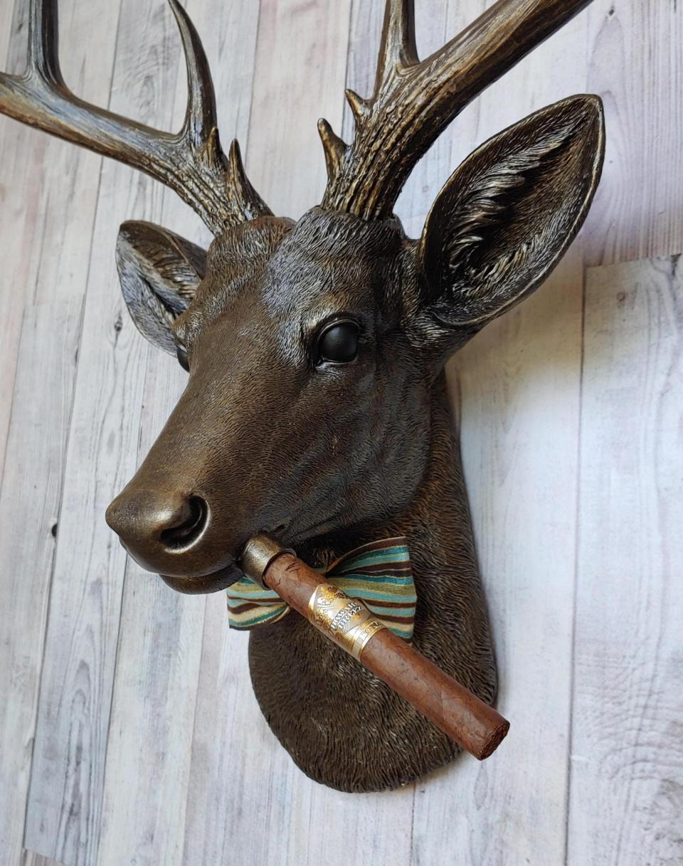 Faux taxidermy deer head painted rubbed bronze with rubbed bronze antlers and gold accents. A cigar is in the deers mouth and a bow tie accents the deers neck.