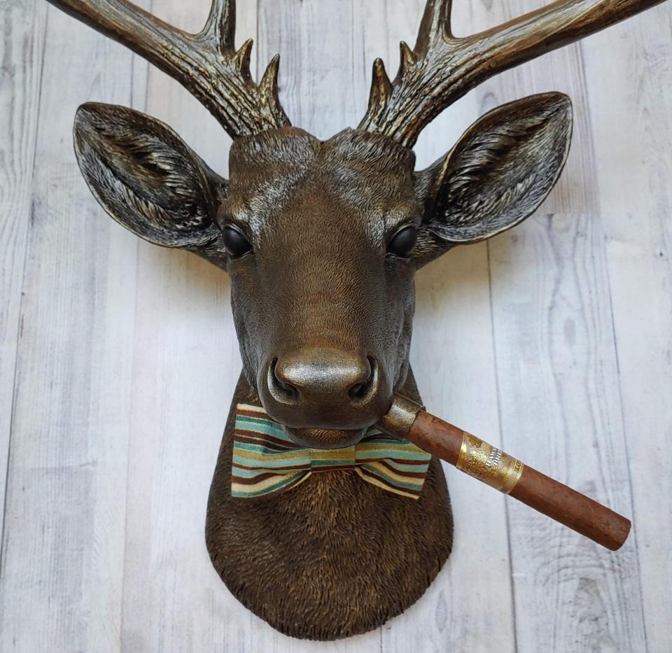 Faux taxidermy deer head painted rubbed bronze with rubbed bronze antlers and gold accents. A cigar is in the deers mouth and a bow tie accents the deers neck.