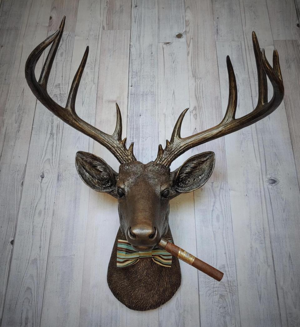 Faux taxidermy deer head painted rubbed bronze with rubbed bronze antlers and gold accents. A cigar is in the deers mouth and a bow tie accents the deers neck.