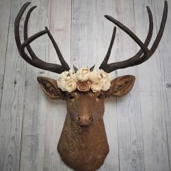 Faux taxidermy deer head that's been painted antique gold with chestnut brown accenting. The antlers are chestnut brown and atop the deers head is a cream floral crown made out of wooden sola flowers.
