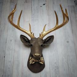 Faux taxidermy deer head, painted chestnut brown with gold accents. The antlers are gold and there is a multi colored brown tonal bow tie on the deers neck.