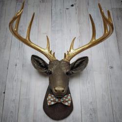 Faux taxidermy deer head. Painted chestnut brown with gold accents and gold antlers. There is a blue and brown polka dot bow tie on the deers neck.