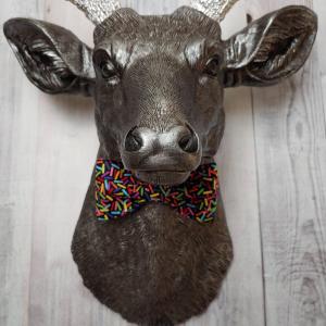 Faux taxidermy deer head painted rubbed bronze with silver accents and silver antlers. The deer has a bow tie on it's neck in black with colorful sprinkles.