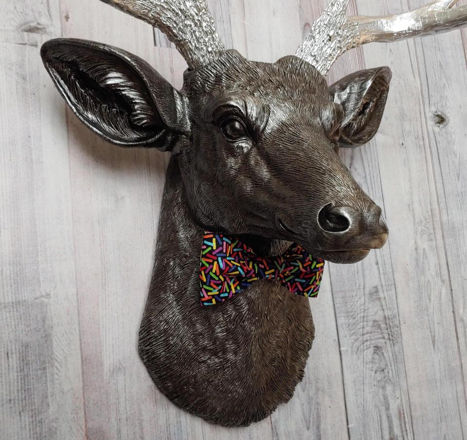 Faux taxidermy deer head painted rubbed bronze with silver accents and silver antlers. The deer has a bow tie on it's neck in black with colorful sprinkles.