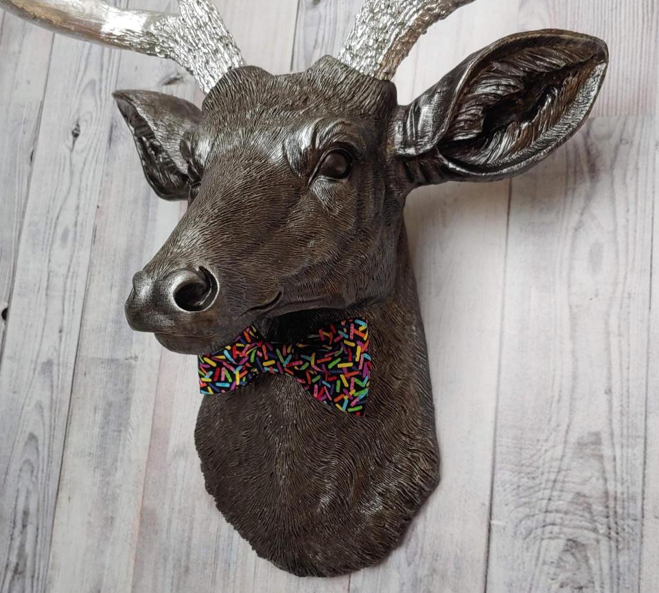 Faux taxidermy deer head painted rubbed bronze with silver accents and silver antlers. The deer has a bow tie on it's neck in black with colorful sprinkles.